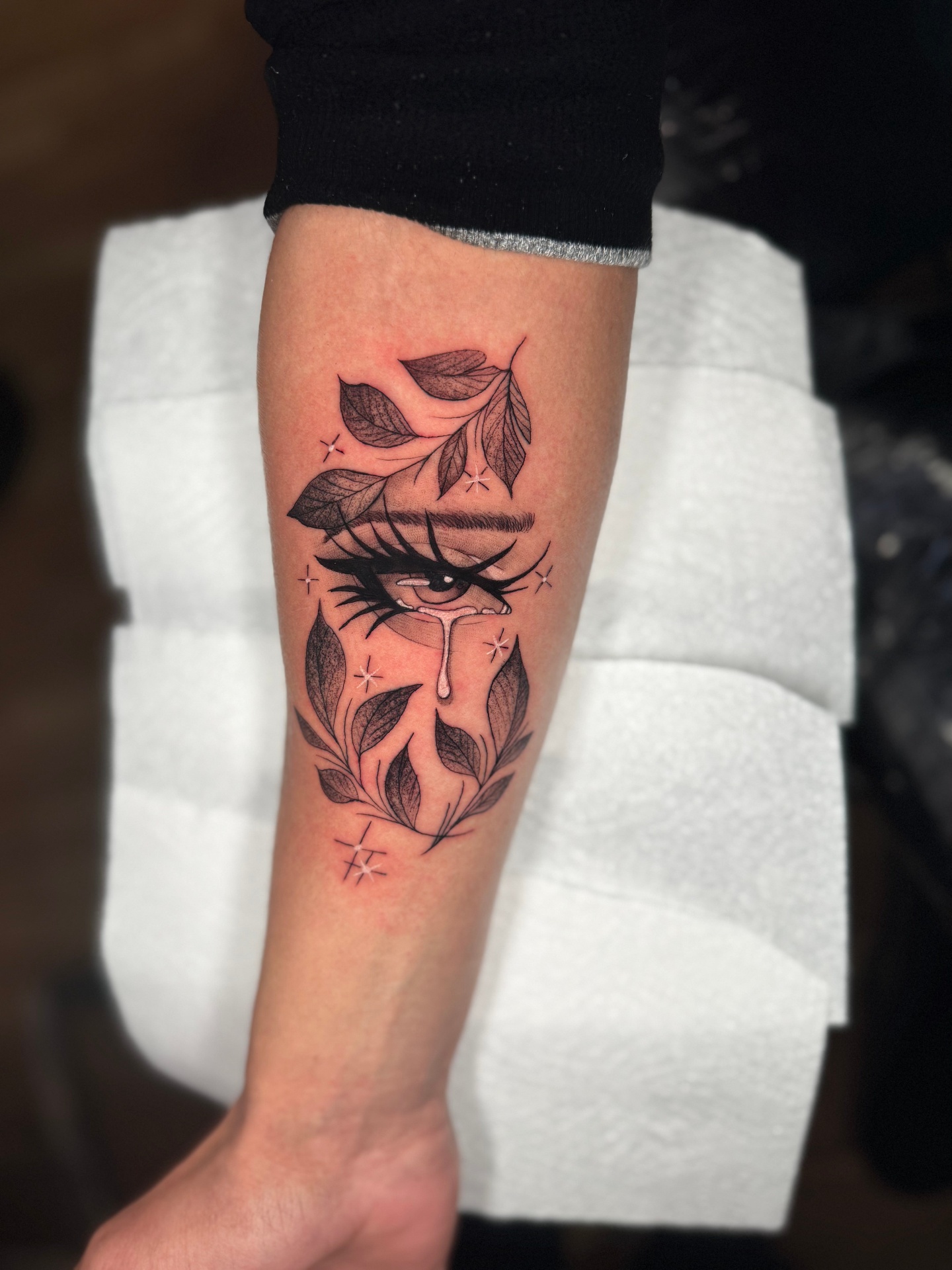 Black and grey tattoo of a crying eye surrounded by leaves on the forearm