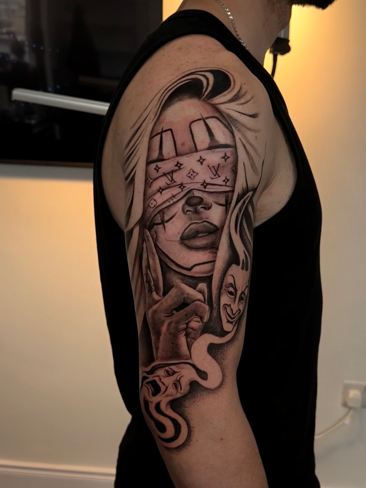 Black and grey chicano-style tattoo of a blindfolded woman with drama masks on the upper arm