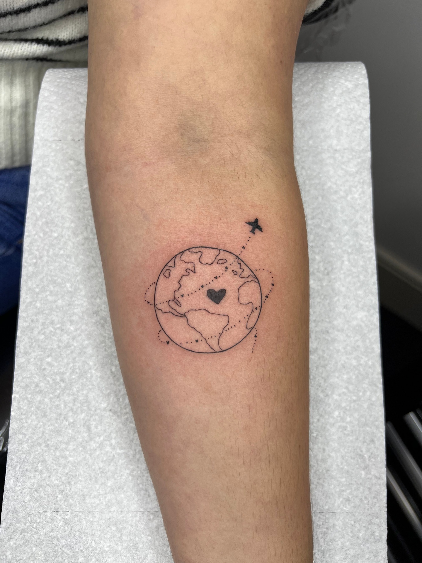 Fine line tattoo of the earth with a plane and heart on the forearm